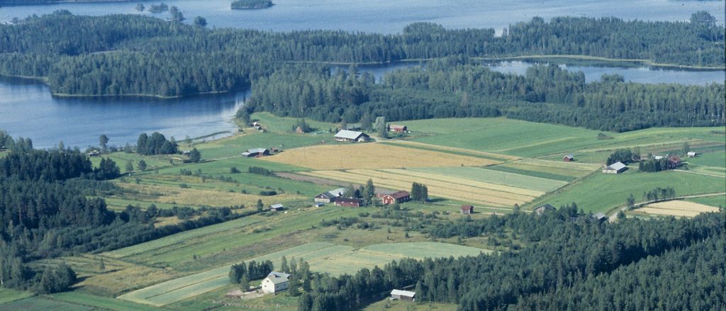 Maatilan rakennuksia peltojen ja metsien keskellä. Taustalla vesistöä.